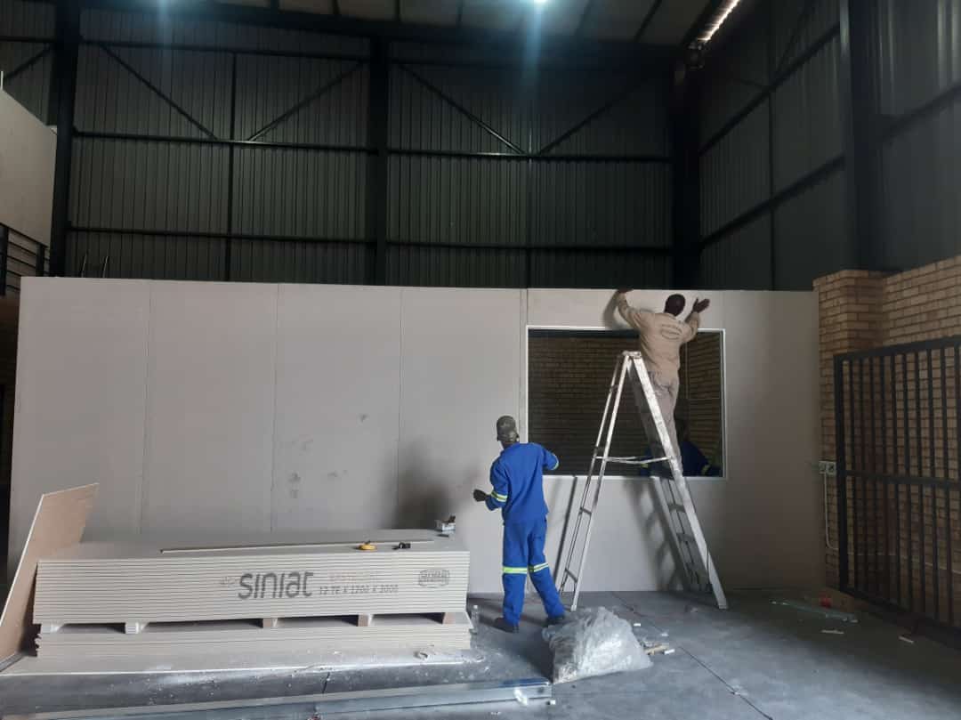 "Two workers installing drywall (Siniat brand) in a large warehouse or commercial space. One worker is on a ladder near a window opening, and the other is on the floor next to a stack of gypsum boards."