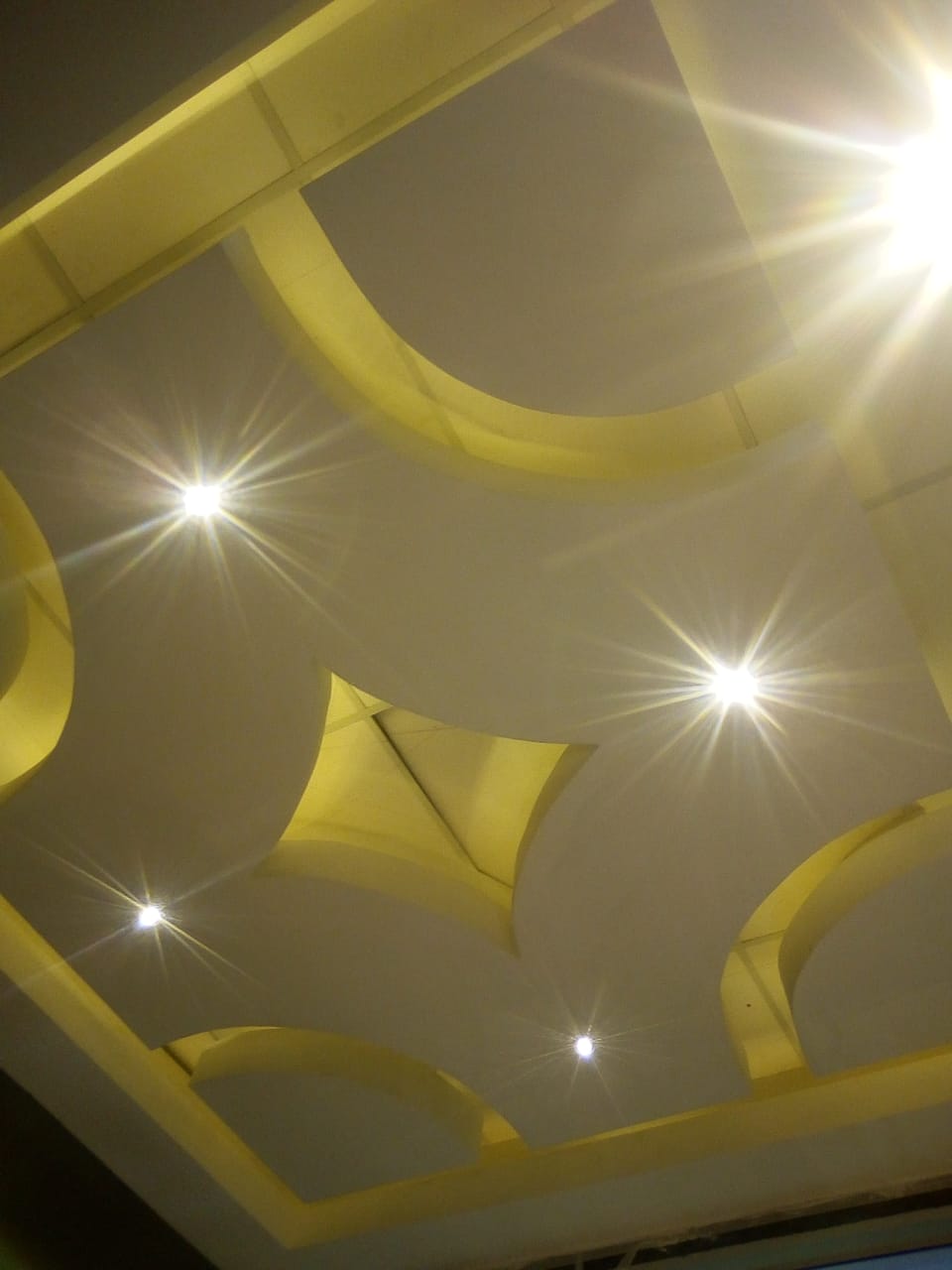 The image shows a modern, stylish drop ceiling installation featuring complex geometric shapes (arches and a central star-like shape) illuminated by warm-toned indirect lighting hidden within the coves. Several bright, star-burst-like spotlights are recessed into the ceiling panels, creating dramatic illumination.